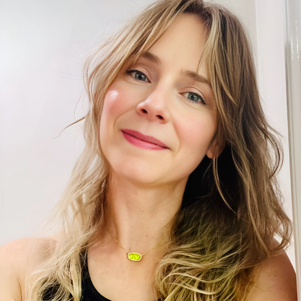 Woman with blonde hair wearing a necklace with a green pendant against a white background